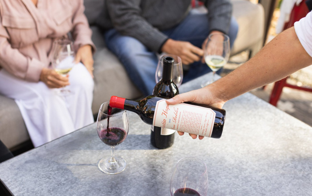 Two people are seated for a wine tasting as someone pours a glass of an Austin Hope wine