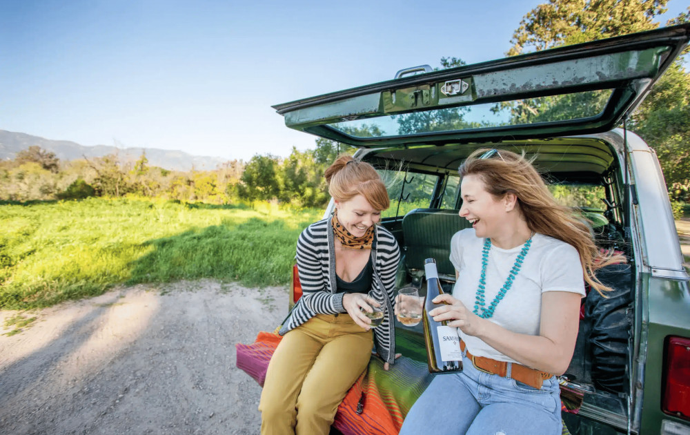 Two women sit in the back of an open SUV sharing a bottle of wine